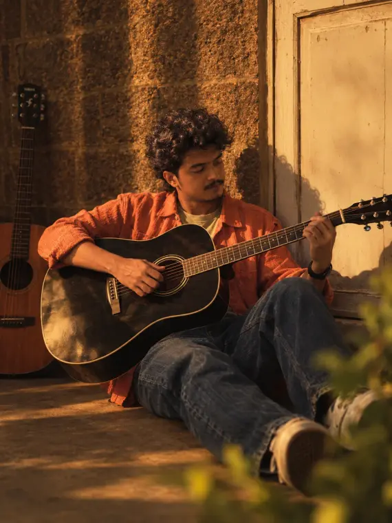Mariappan sitting against a stone wall in golden-hour light, playing a black acoustic guitar, a second guitar leaning beside him