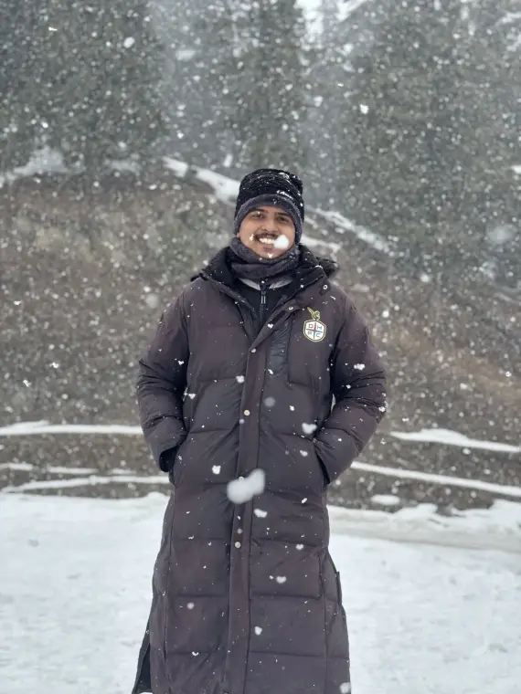 Mariappan grinning in a long puffer coat and beanie as snow falls thick around pine trees