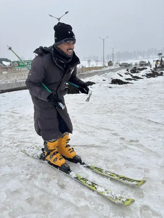 Mariappan on skis in yellow boots, gripping the poles on a snowy slope with a misty valley behind
