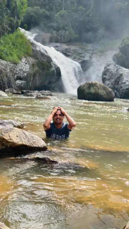 Mariappan neck-deep in the river pool below a waterfall, hands on his head, smiling