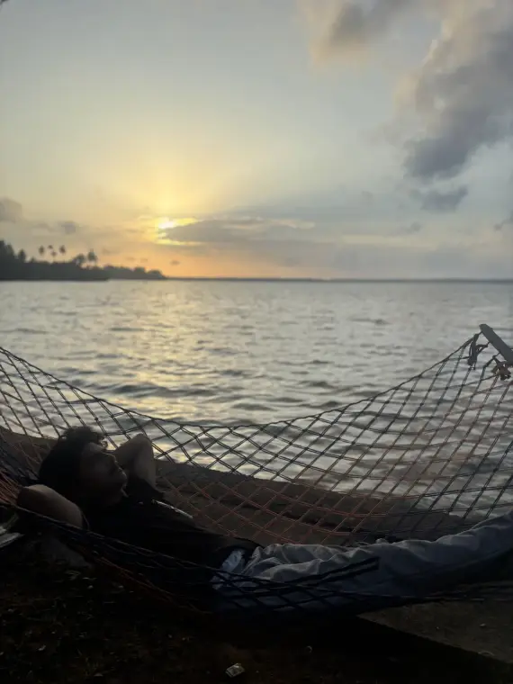 Mariappan lying back in a rope hammock by the water as the sun sets over a palm-lined shore