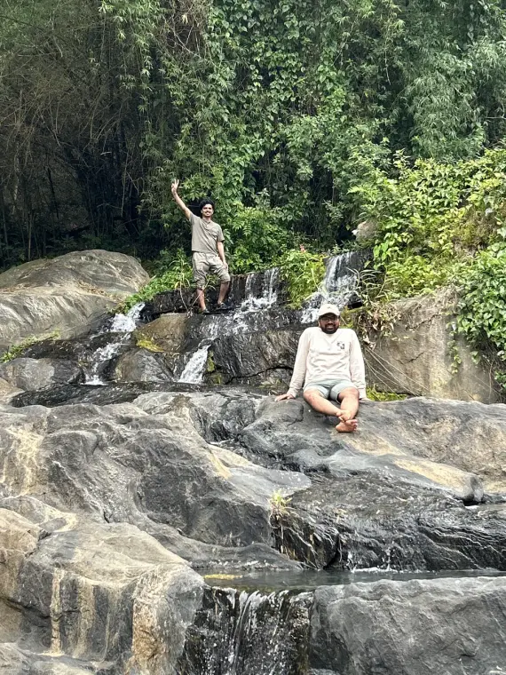 Two friends on the rocks of a small forest waterfall, one waving from the top, one sitting below