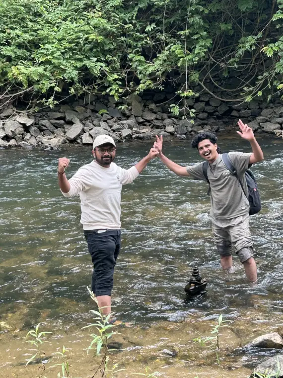 Mariappan and a friend knee-deep in a stream, cheering beside a little stack of balanced stones